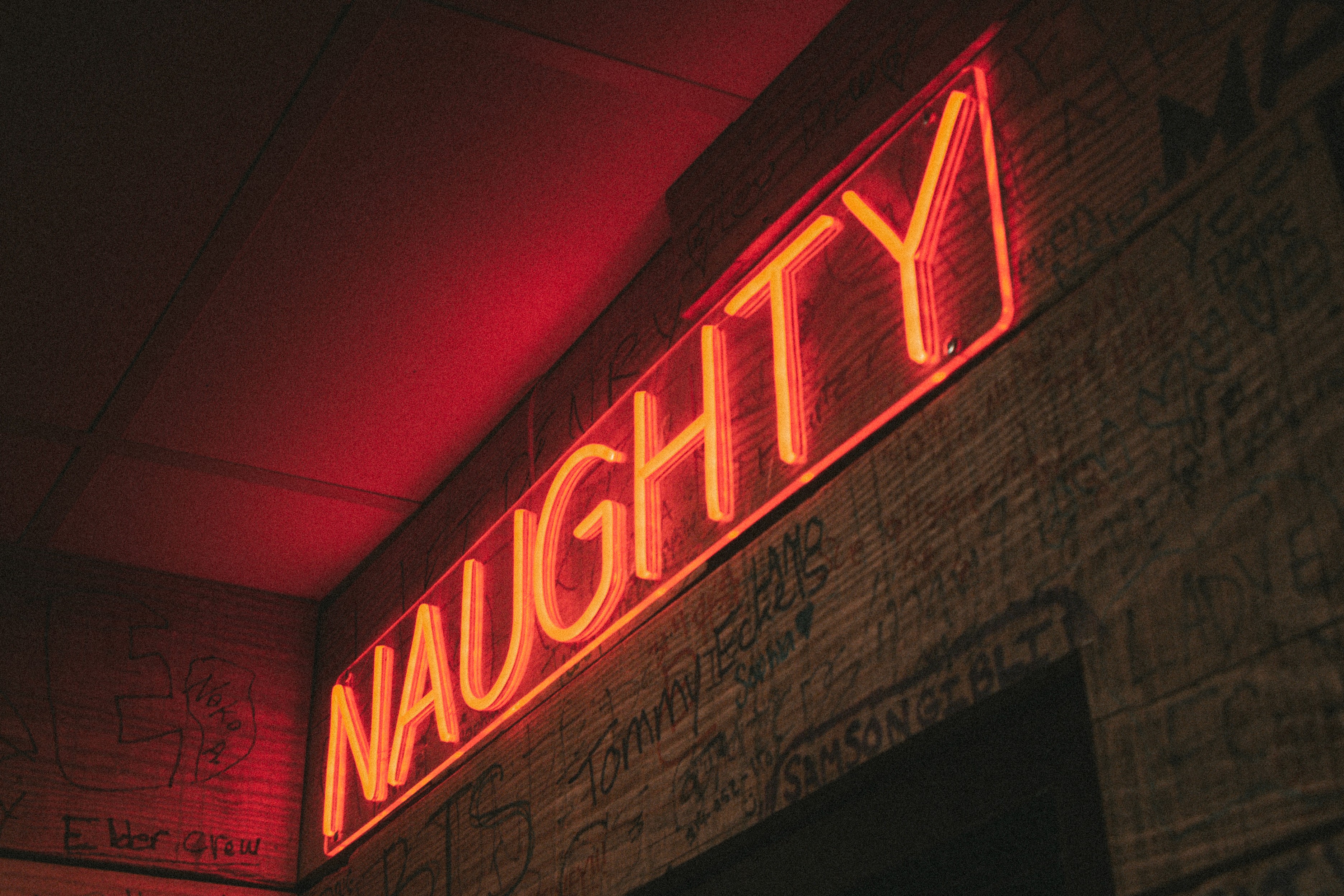 A red neon NAUGHTY sign glowing over a wall covered in patron signatures